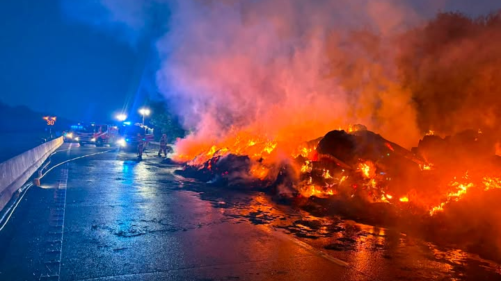 A huge pile of material on fire on a motorway at night. Firefighters and fire engines with blue lights can be seen in the background.