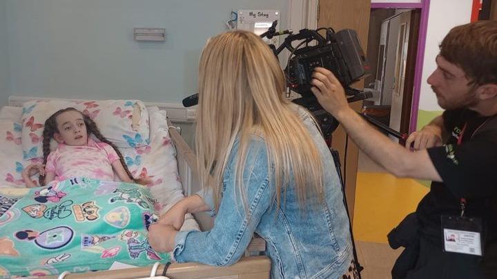 A cameraman is filming a young girl as she is being interviewed in her bedroom at Derian House. She is wearing pink clothes and is lying in bed. The cameraman and another woman are standing at the corner of her bedroom.