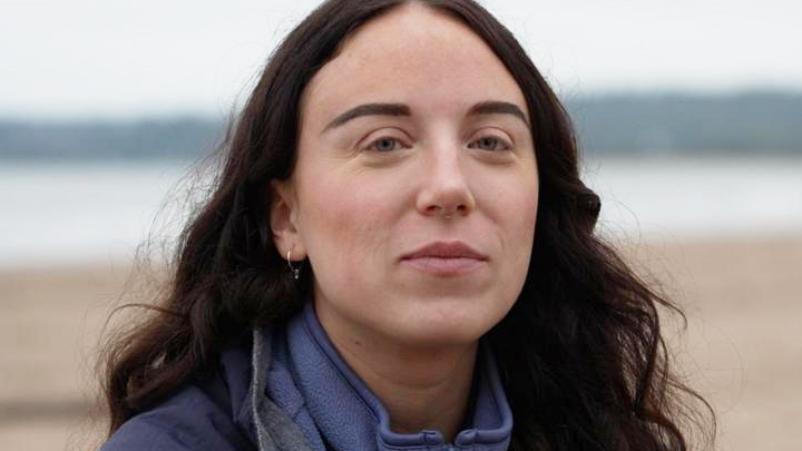 Amy Rae smiling at the camera with a beach in the background. She is wearing a blue sweatshirt. 