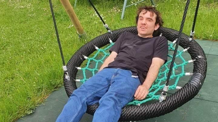 Declan Morrison sitting on a large netted, basket style swing. He is mid swing and is looking at the camera. He is wearing a black T-shirt and blue jeans. He has both of his hands by his side