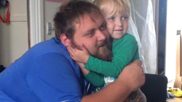 A young boy with blonde hair, wearing a green long sleeve top, cuddles a bearded man with brown hair, who is wearing a bright blue t-shirt.