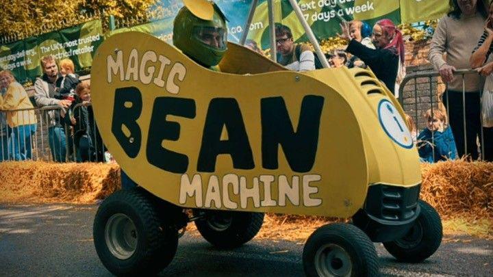 A man in small vehicle that has four wheels and has been decorated to look like a bean. He is taking part in a go-kart race. The vehicle is yellow and and reads "magic bean machine" on the side. Crowds are gathered behind the metal fences and hay bales to watch.