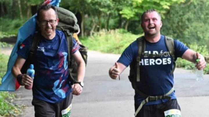 Jim Brace and Craig Smith running. Craig has a help for heroes t-shirt on and he is giving a thumbs up. Jim has a drink bottle in his hand and a backpack on. 

