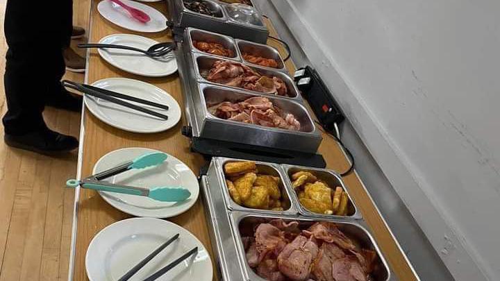 A picture of several containers that sit on a long table and are filled with various breakfast foods including bacon, hash browns and more. Plates with tongs rest in front of the containers.