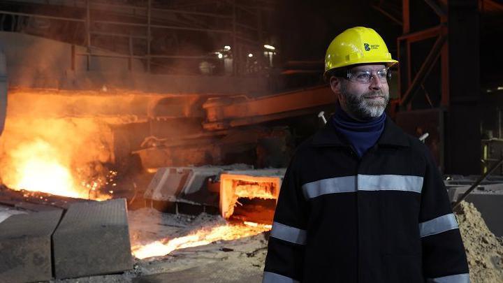 Joanthan Reynolds poses in front of molten metal at a blast furnace at British Steel's Scunthorpe plant. He is wearing a heavy black protective coat, goggles and a yellow hard hat