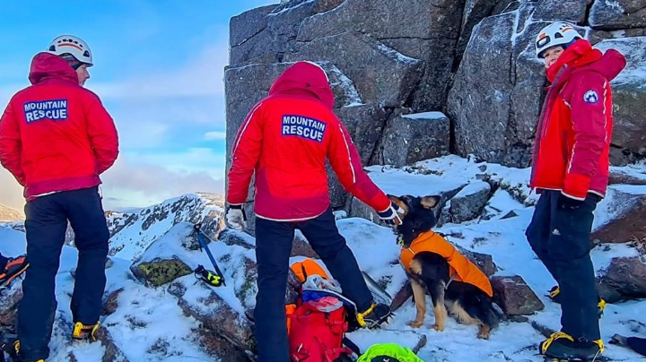 Three people with mountain rescue coats on a snowy outcrop with the dog Bramble wearing an orange coat seated at their feet 