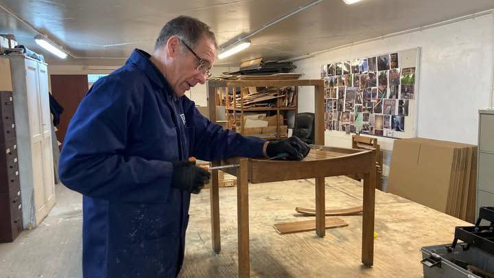 A man in blue overalls holding a screwdriver to the seat of a wooden chair. He has black greying hair and is wearing glasses. He is standing in what appears to be a store room, the chair placed on a large wooden table, with assorted items of furniture and off-cuts on shelves around him.