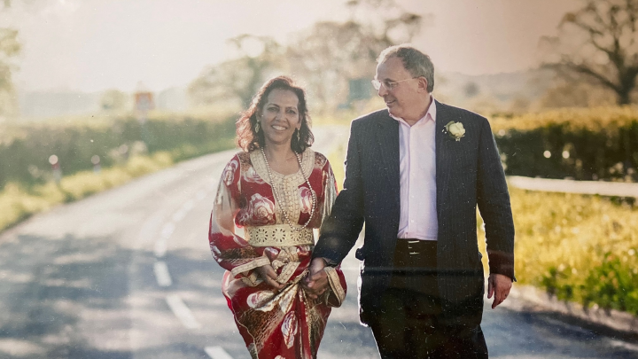 Andrew and his wife Zoubida on their wedding day walking down a country road. He is wearing a navy stripped suit and she is wearing a red and gold full-length dress