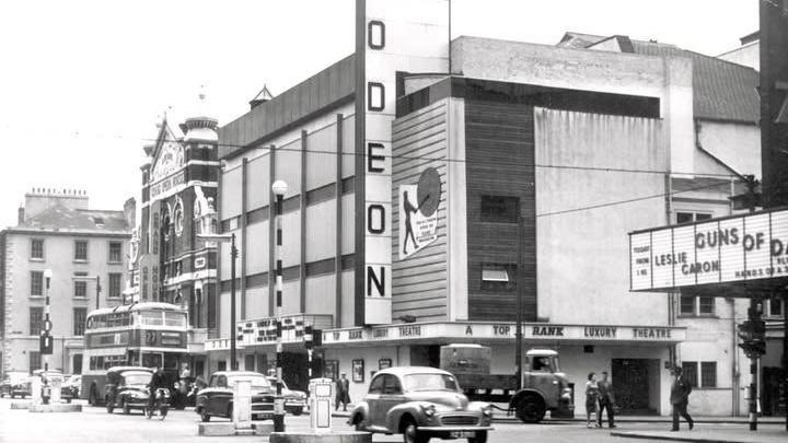 A black and white image of a busy street with old fashioned cars and a concrete theatre with a ODEON sign at the front