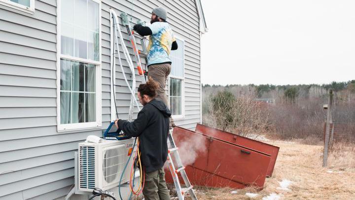 Workmen fit a heat pump to the to the clapboard exterior of a house.