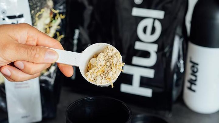 A person holding a scooper with Huel powder in it. They are holding it over a black cup and there is a Huel bag in the background