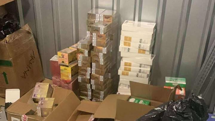 Brown and white boxes of illegal vapes stacked on top of each other within a storage unit. Some smaller boxes sit in larger brown boxes. A black bin back can be seen to the right of the image.