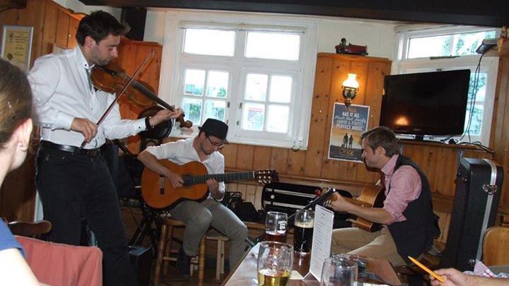 Three men are playing instruments in a pub. Two people play guitars and another plays a violin. People are watching the musicians while drinking at their tables.