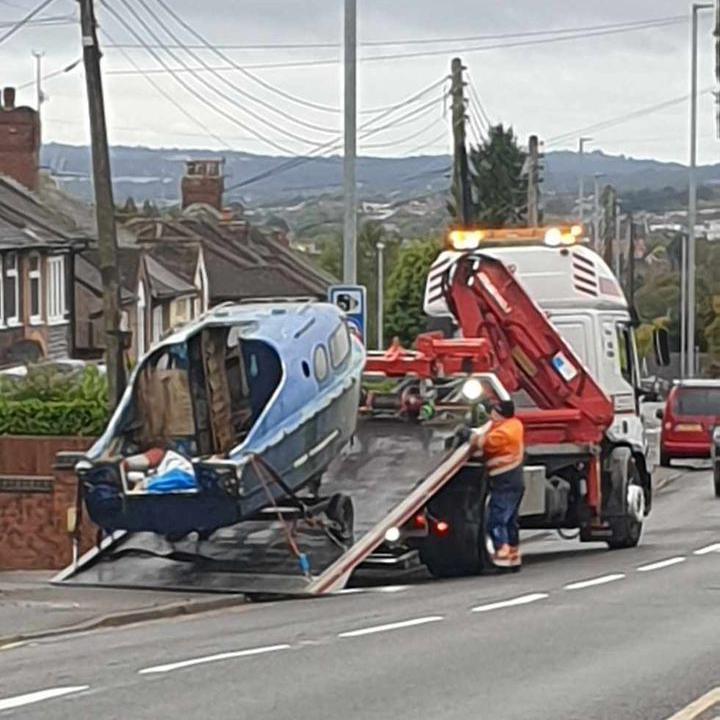 A small blue boat is being loaded onto the back of a flatbed truck, with a man in an orange high visibility jacket stood next to it.