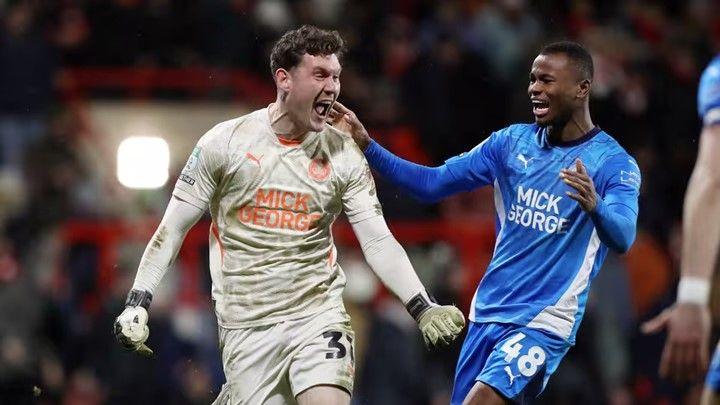 Peterborough United goalkeeper Jed Steer, wearing a long sleeve cream top and shorts, runs with arms outspread and his mouth agape as he celebrates a penalty save.