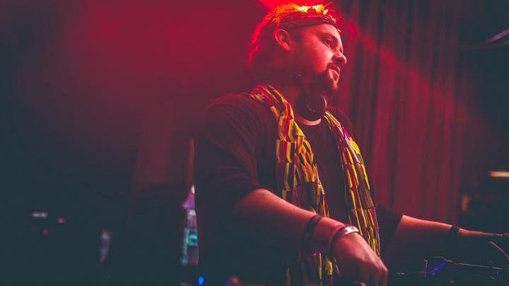 Dan Wild in a yellow and red scarf DJing. The lighting is red, making most of the image red. He has headphones round his neck.