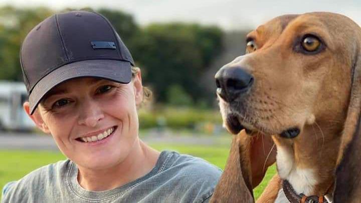 A young woman sits next to a large dog, with both of their heads level. She is wearing a baseball cap and a grey T-shirt and is smiling at the camera. The brown dog, with floppy ears, stares straight ahead. 