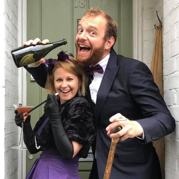 Tom Price, in a tuxedo, holds a bottle of fizz and a walking stick, while Beth, in a purple evening dress and fur stole, poses with a full Martini glass and cigarette holder on their doorstep.