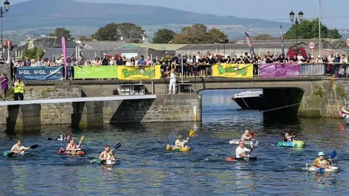 People in tin baths in Castletown Harbour, with hundreds of spectators watching from a road bridge across the harbour which has banners on it.
