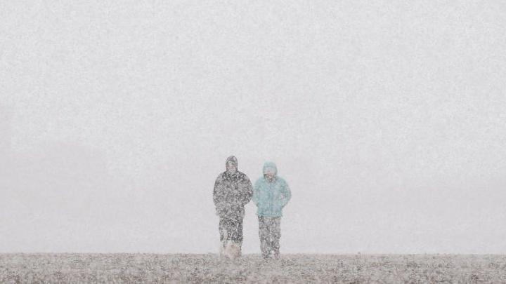 Two people in thick coats walk through falling snow across a flat landscape