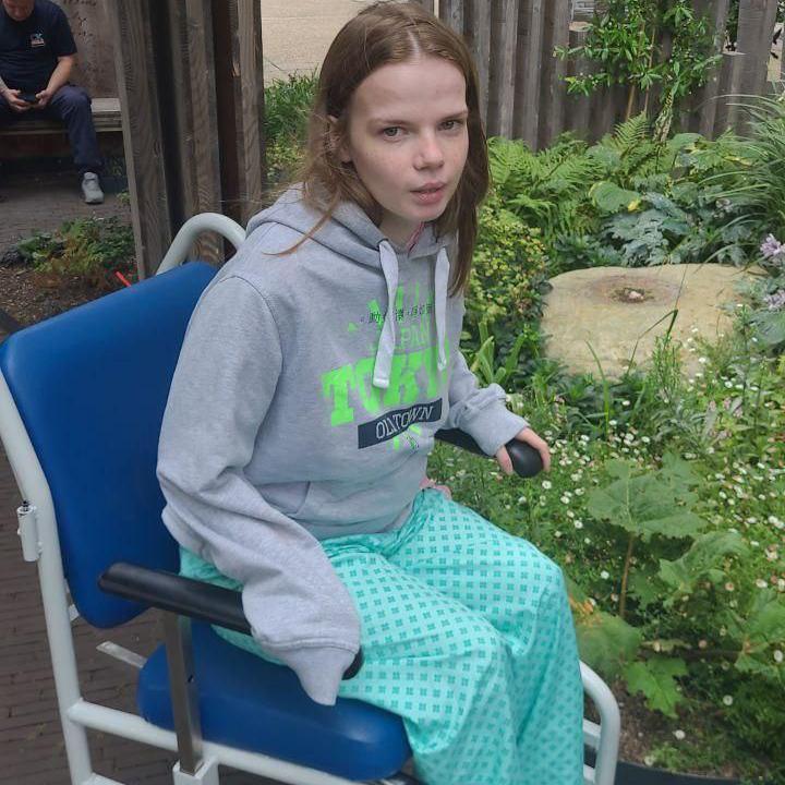 Anastasija is seen in a wheelchair outdoors wearing a grey hoodie and hospital gown. She is positioned near greenery.