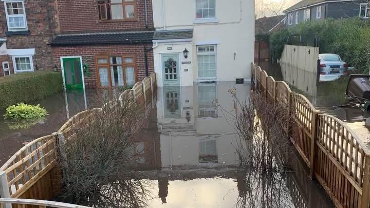 A view of the rear of a row of terraced houses which all have rear gardens submerged in brown, muddy flood water