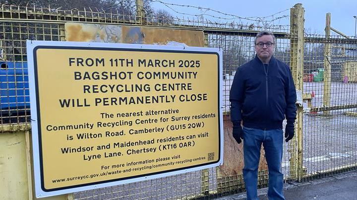 A man wearing a black jacket and black gloves stood to the right of a big yellow sign. The yellow sign reads: "From 11th March 2025 Bagshot Community Recycling Centre will permanently close / The nearest alternative Community Recycling Centre for Surrey residents is Wilton Road, Camberley (GU15 2QW) / Windsor and Maidenhead residents can visit Lyne Lane, Chertsey (KT16 0AR)"