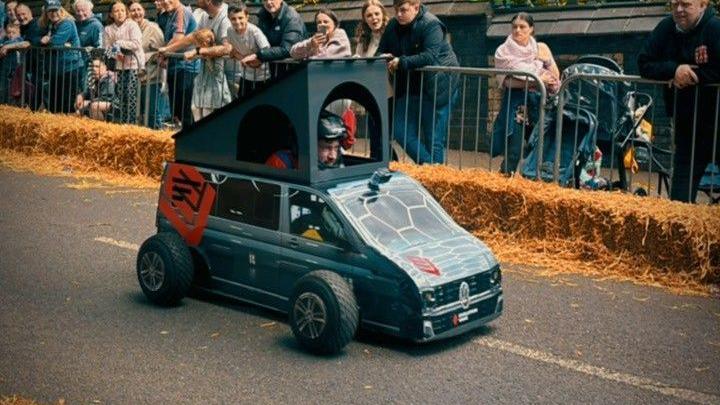 A man in small black vehicle that is taking part in a go-kart race. The vehicle has been made to look like a black transit van with a camping tent on top. The crowd are standing back off the road and are behind a metal fence and hay bales. 