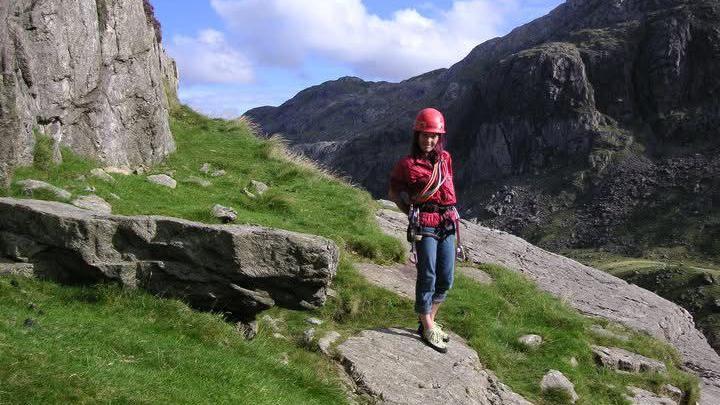 A woman wearing a pink helmet, pink jacket and jeans stands on a rock on the grass in the middle of a rocky and mountainous terrain.