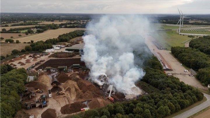 An aerial image taken by a drone looks down on a commercial wood site with big piles of bark, some of which are giving off large clouds of smoke from a fire, with the site shown surrounded by fields, one of which includes solar panels and two large windmills.