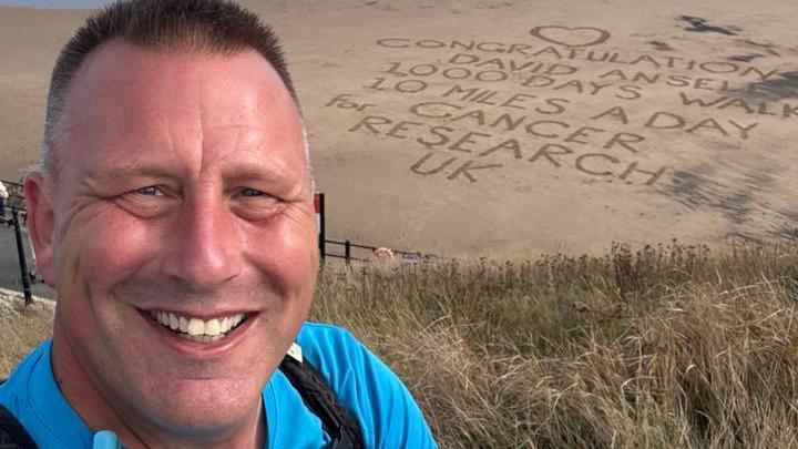 David Ansell smiling into the camera. He has short, brown hair which is grey on the sides. He is wearing a bright blue top. There is a beach behind him and words have been written into the sand along with a heart. It reads: 'Congratulations David Ansell 1000 Days walking 10 miles a day for Cancer Research UK.'