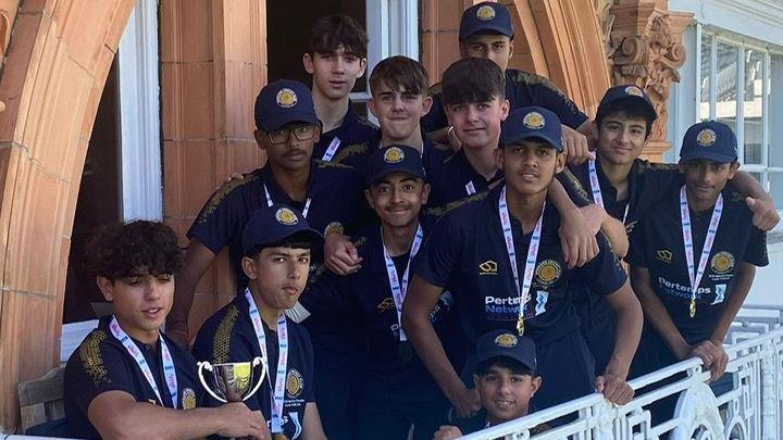 Twelve members of an U15 cricket team, dressed in navy, on the balcony at Lord's. They are all wearing medals and one member is carrying a cup.