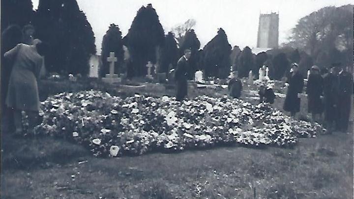 A black and white image of lots of flowers over several graves. There are mourners dressed in black at the side of the graves.