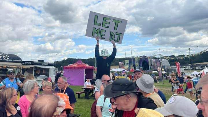 A young boy wearing a green and white bucket hat, a black sweatshirt and light grey shorts. He's sitting on the shoulders of a bald man and holding up a large white sign saying 'let me DJ' on in green letters. He's at a festival in the daytime, and there are crowds of people, tents and bunting all around him.