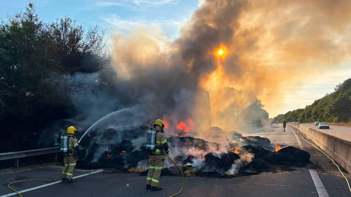 Two firefighters with hoses spray water on a pile of scorched material next to a lorry on a motorway.