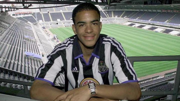 Kieron Dyer wearing the black and white striped Newcastle United jersey. He is smiling at the camera while positioned high up at St James' Park stadium, which can be seen in the background with its green grass pitch and surrounding stands.