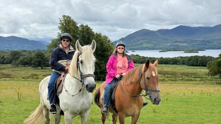 Two people sat on horses, looking at the camera. On the left is a man wearing blue denim jeans and a beige and navy coat. He is on a white horse. On the right is a woman wearing a salmon-pink t-shirt and a pink coat. She is on a brown horse. Both people are wearing sunglasses and riding helmets. They are in an empty field. In the distance, there are dozens of trees, a body of water, and some hills.