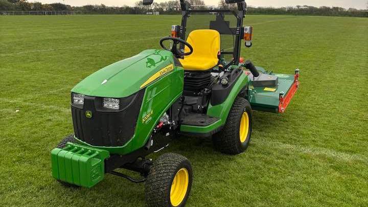 A picture of a green husqvarna ride-on mower. The mower is on what looks like a football pitch and there are trees and bushes in the background.