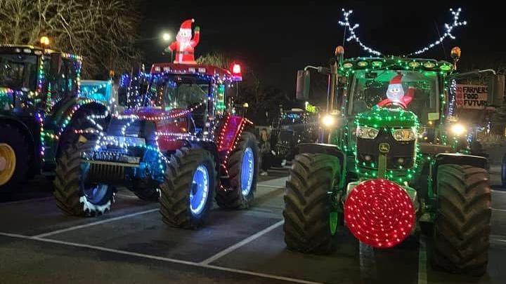 A blue and green tractor at the centre front of the photo, both decorated with Christmas lights and Santa effigies, one of which is behind the wheel in the cab on the right, other tractors can be see in the background of a car park.