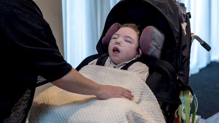 A person is gently adjusting a cream-colored blanket over a child seated in a black wheelchair. The wheelchair has a colorful patterned side panel. The background features white drapes and a striped carpet, suggesting an indoor event setting.