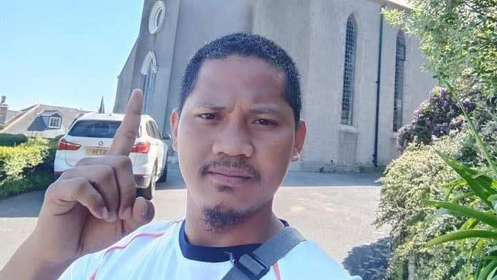 George Michael Monte De Ramos Castrudes wearing a white t-shirt and pointing upwards while standing in front of a church. He had short, black hair and a moustache and goatee beard. 