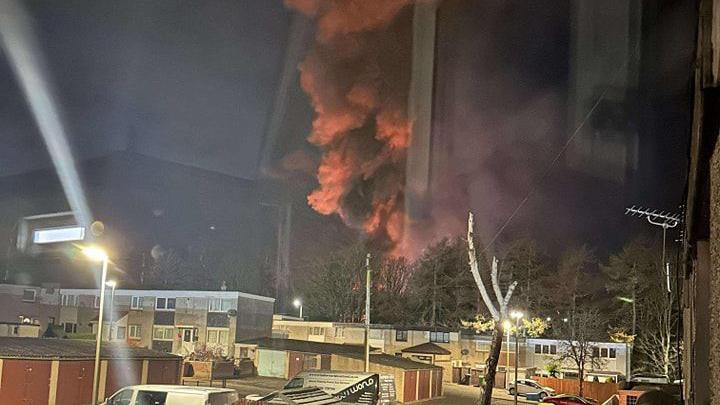 Smoke rising from behind a house during a fire at a paper mill in Fife. The smoke is grey and orange coloured against a dark, overcast background. Several houses and garages can be seen in the foreground.