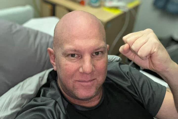 Adam Claxton lies in a hospital bed with a fist raised in the air while he looks at the camera. He wears a black t-shirt with green sleeves, while a tattoo is visible on his arm.