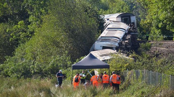 Three derailed train carriages lie on their sides on a railway embankment near Lewes in East Sussex. Rail workers in orange high-visibility clothing and a police officer are beside the track as investigators examine the scene. One carriage doorway is visible, showing damage where the train separated during the derailment.