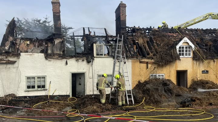 Two fire fighters are holding a ladder against a white painted property. There are two properties which are both next to each other with roof damage following a fire. The roof on each property has been burnt and you are able to see into the properties due to the amount of damage. 