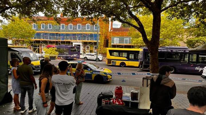 Pedestrians watch on from the pavement following the bus crash. The crashed bus can be seen in the distance behind a cordon with police cars parked up as well as other buses.
