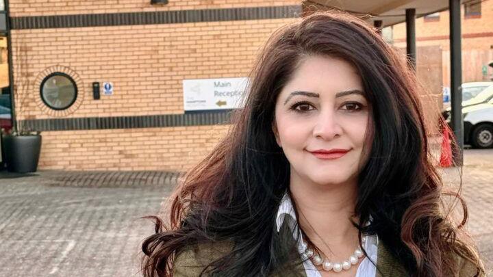 Dr Shabina Qayyum stands in the car park of a doctor's practice. She has dark flowing hair and is wearing s breen jacket, pearls and a shirt.