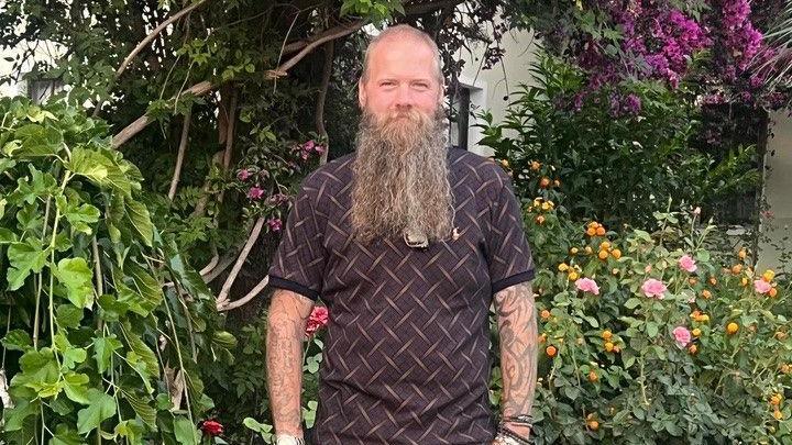 Mr Hammond smiles at the camera in front of bushes and flowers. He has a long beard and wears a patterned polo top.