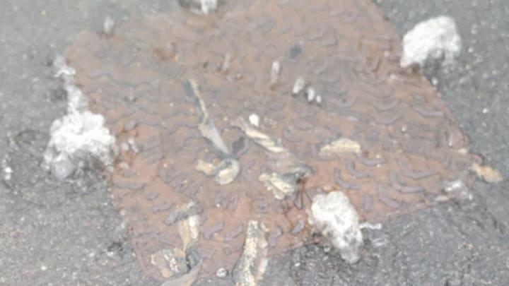 A rusted brown manhole cover, covered in water with white bits of paper strewn around it.