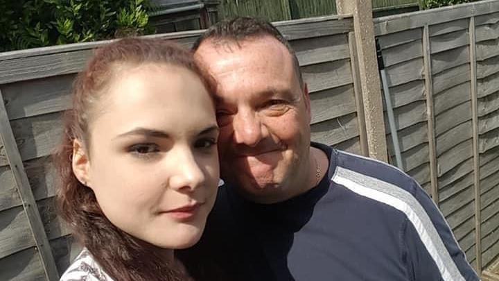 A young woman with brown, curly hair tied back poses for a selfie next to her father. They are outside with the sun shining on their faces. Brown garden fence panels can be seen in the background. They are both smiling and their heads are touching.